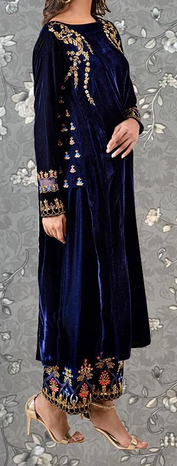 A woman wearing a long, navy blue velvet dress with gold embroidery and floral patterns, standing against a gray floral background.