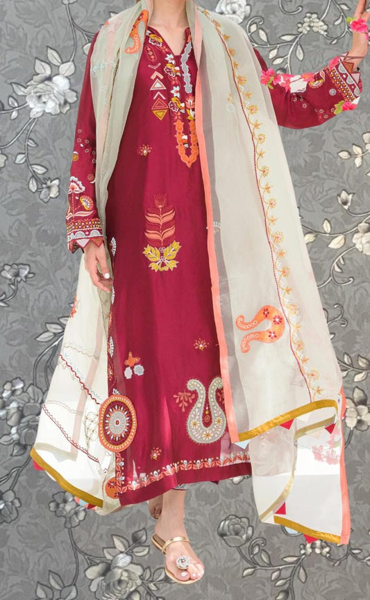 A person wearing a red and gold dress with intricate embroidery and patterns, standing against a gray floral background.