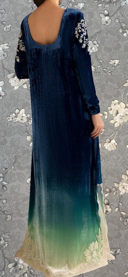 a woman wearing a long, blue velvet dress with floral embroidery on the sleeves and skirt. The dress has a high neckline and long sleeves, giving it a formal and elegant appearance.