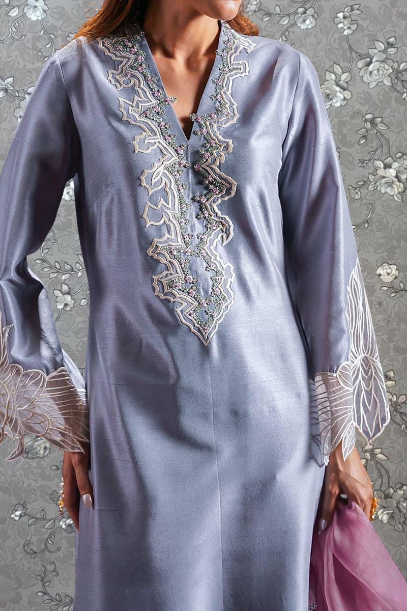 A woman wearing a long, gray dress with intricate embroidery and lace sleeves, standing against a floral background.