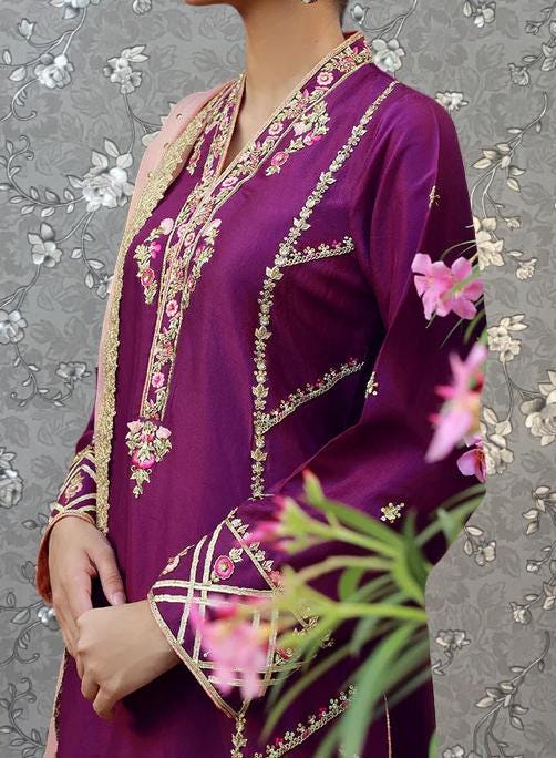 A person wearing a purple dress with intricate embroidery and floral patterns, standing in front of a floral patterned background.