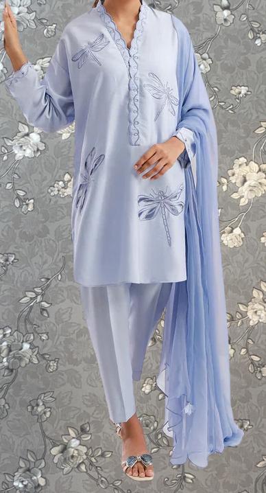 A person wearing a light blue dress with a pattern of blue dragonflies on it, standing against a gray floral background.