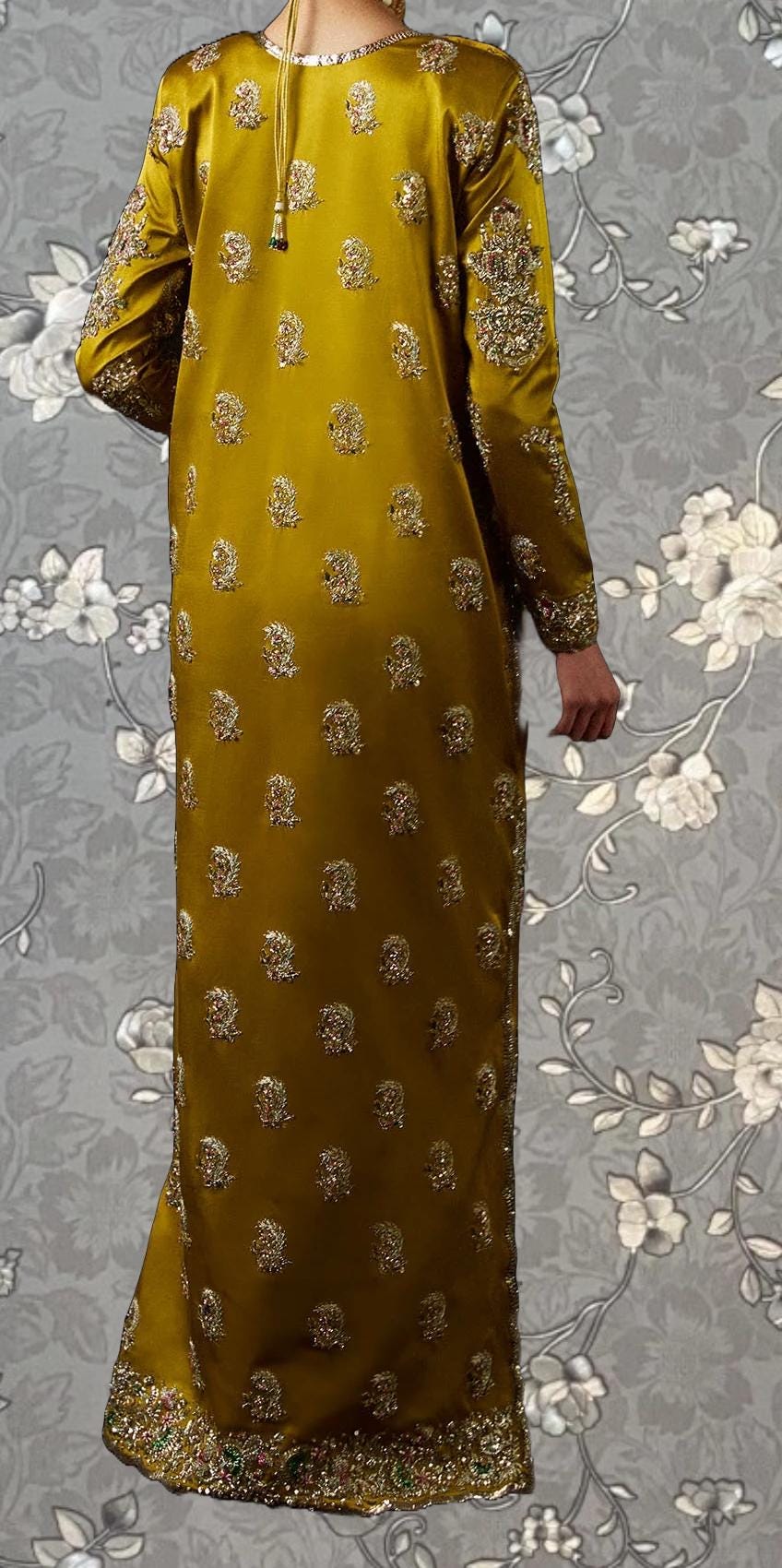 a long, gold-colored dress with intricate embroidery and floral patterns. The dress has a high neckline and long sleeves, giving it a formal and elegant appearance. The background is a gray floral pattern, which complements the gold color of the dress.