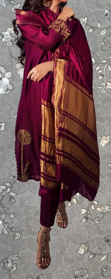 A woman wearing a vibrant purple and gold dress with intricate embroidery, standing against a gray floral background.