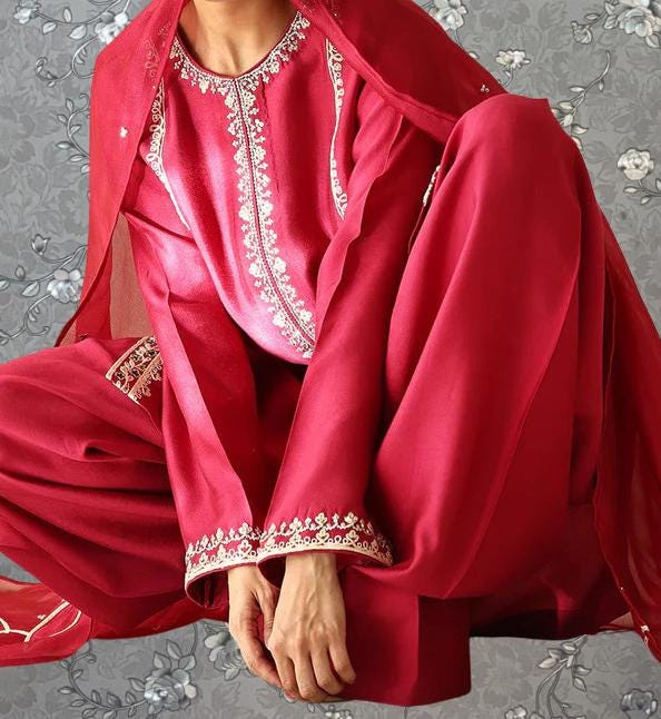 A person wearing a red and gold outfit with intricate embroidery, sitting on a gray floral background.