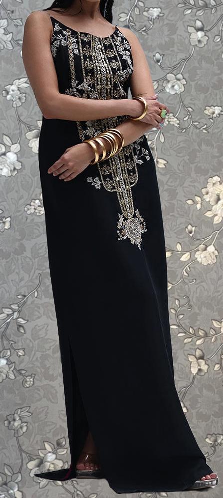 A woman wearing a long black dress with gold embroidery stands against a gray floral background.