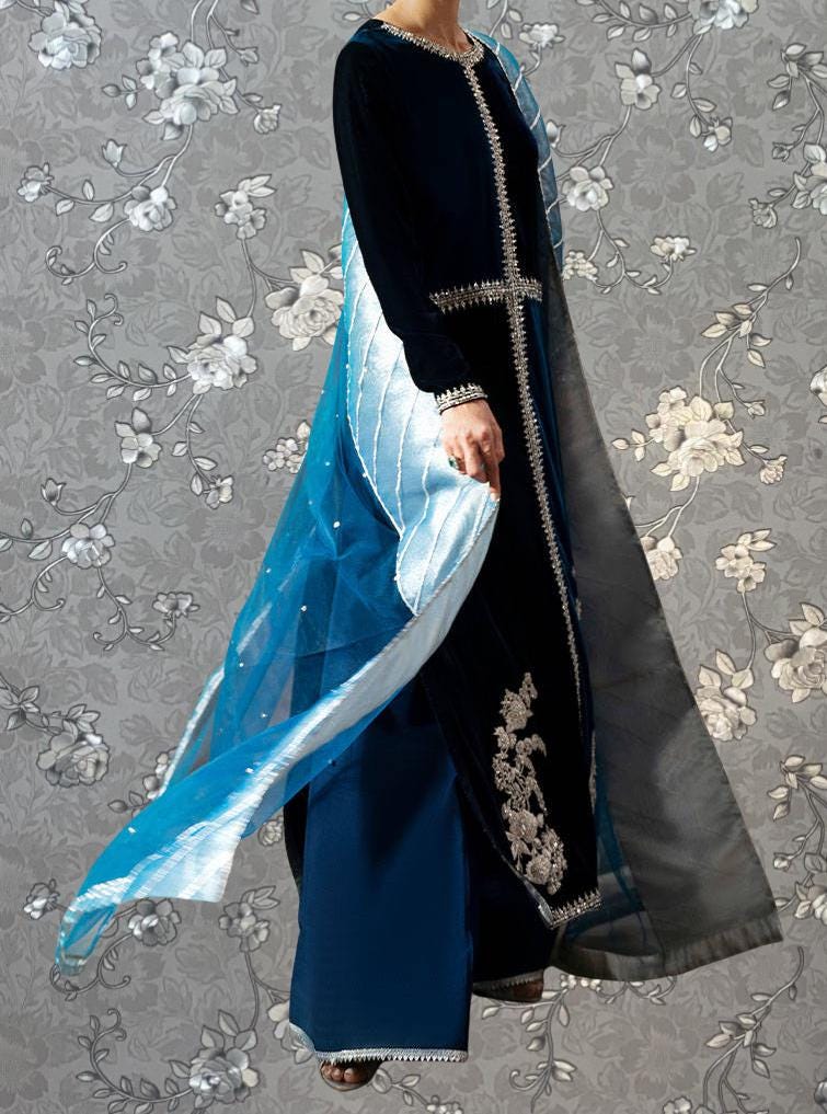 A person wearing a long black dress with blue and white accents, standing against a gray floral background.