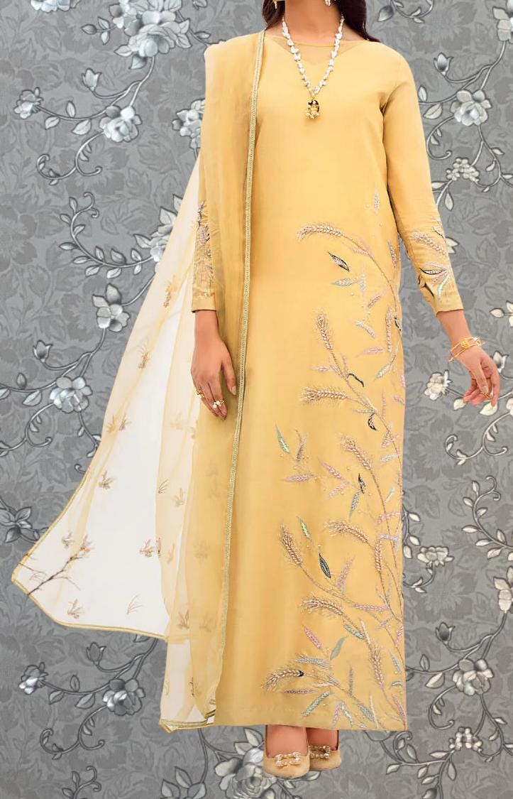 A woman wearing a long, yellow dress with intricate floral embroidery stands against a gray floral background.