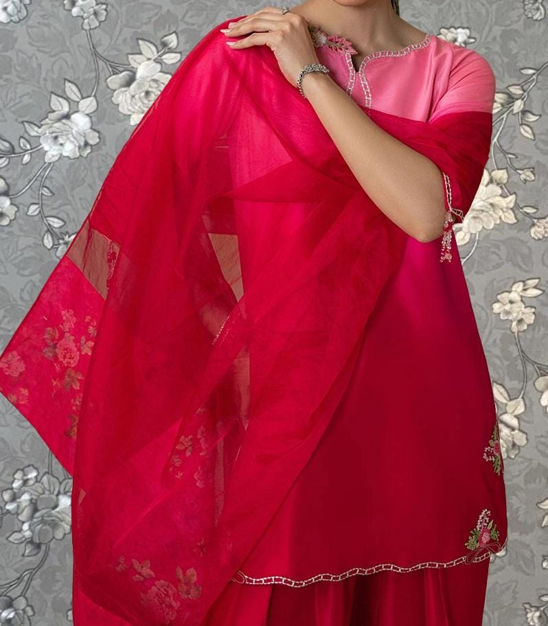 A woman wearing a vibrant red dress with a pink top stands against a gray floral background.