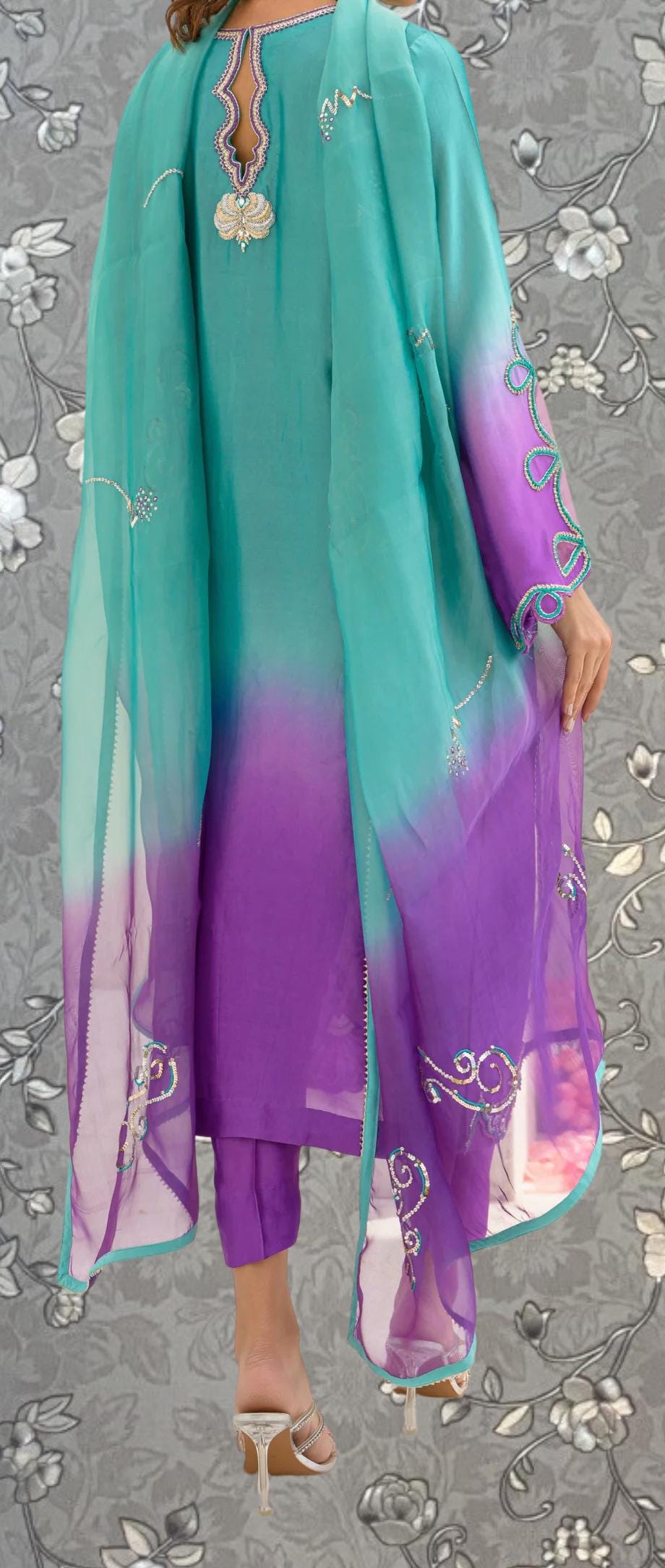 a person wearing a colorful, patterned dress with a turquoise and purple color scheme. The dress features intricate embroidery and designs, and the person is standing against a floral background.