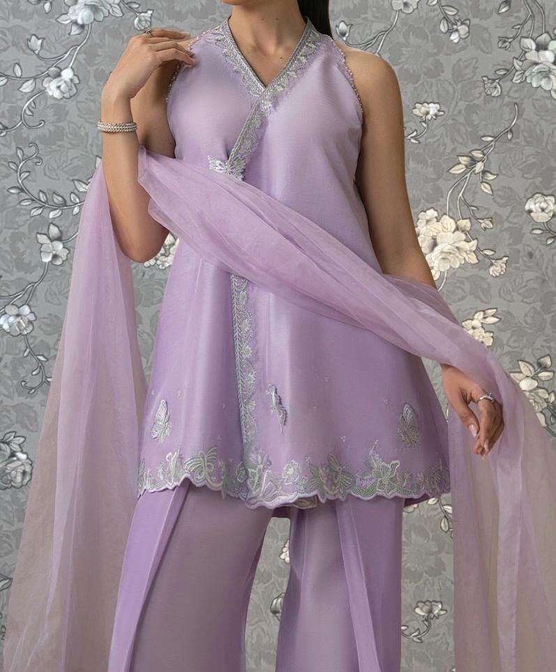 A woman wearing a lavender dress with a floral pattern and a matching shawl stands against a gray floral background.