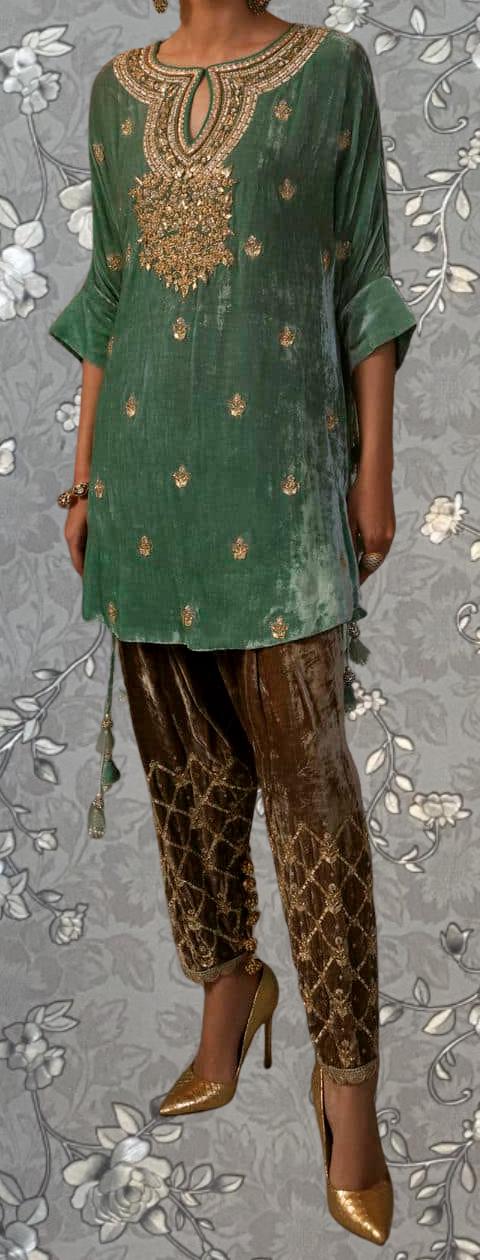 A person wearing a green velvet top with gold embroidery and gold pants, standing against a gray floral background.