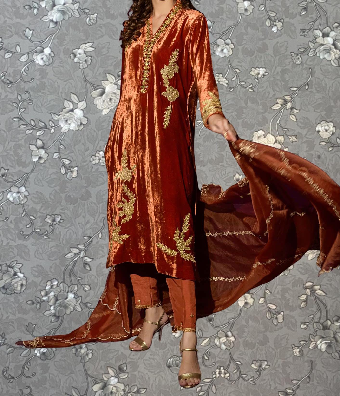 A woman wearing a long, red velvet dress with gold embroidery poses against a gray floral background.
