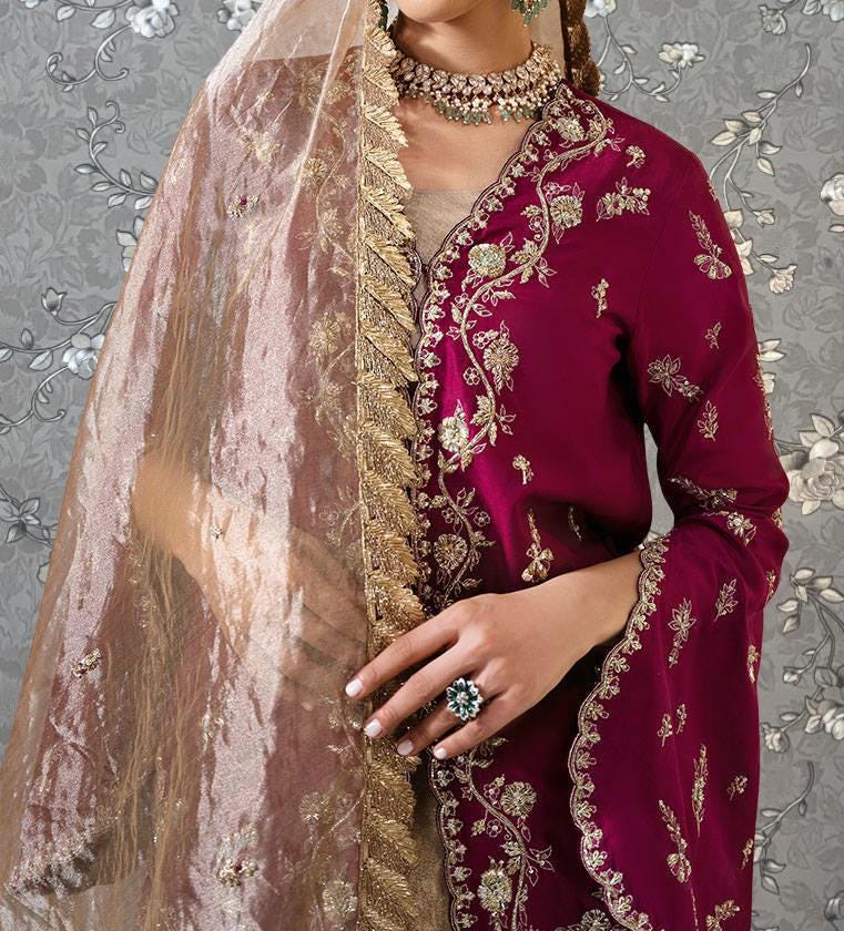 A woman wearing a vibrant red and gold embroidered dress with a matching veil, posing against a gray floral background.