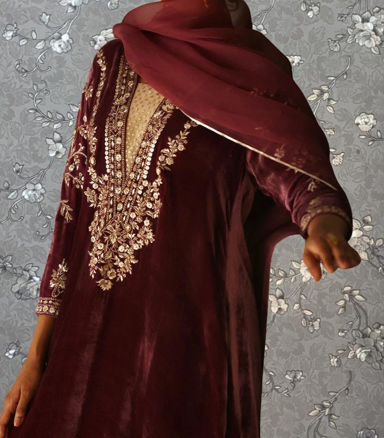 A person wearing a maroon velvet dress with gold embroidery and a matching red scarf stands against a gray floral background.