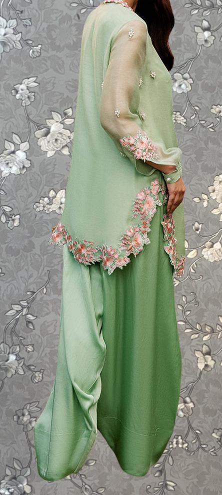 A woman wearing a green dress with pink floral accents, standing against a gray floral background.