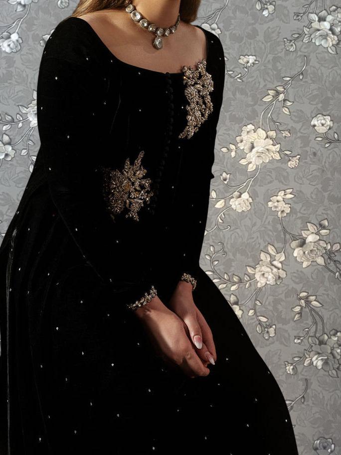 A woman wearing a black dress with gold embellishments stands against a gray floral background.
