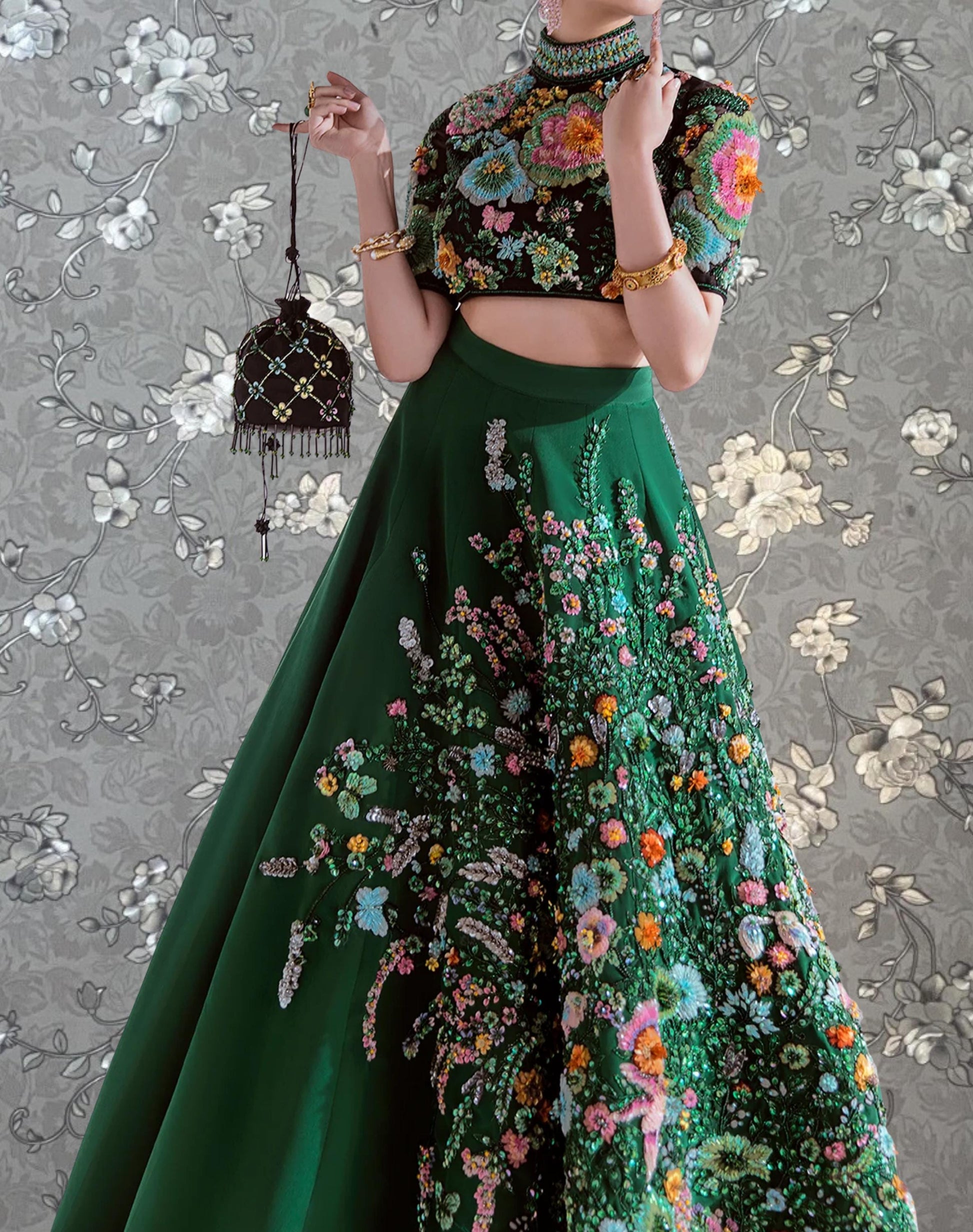 A woman wearing a green and pink floral dress with a high neckline and a black purse is posing against a gray floral background.