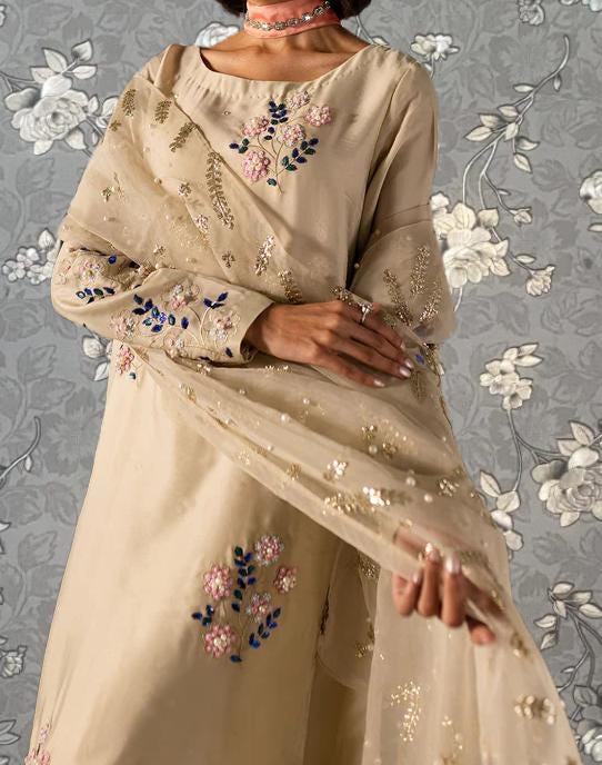 A woman wearing a beige dress with intricate embroidered designs, including flowers and leaves, stands against a gray floral background.