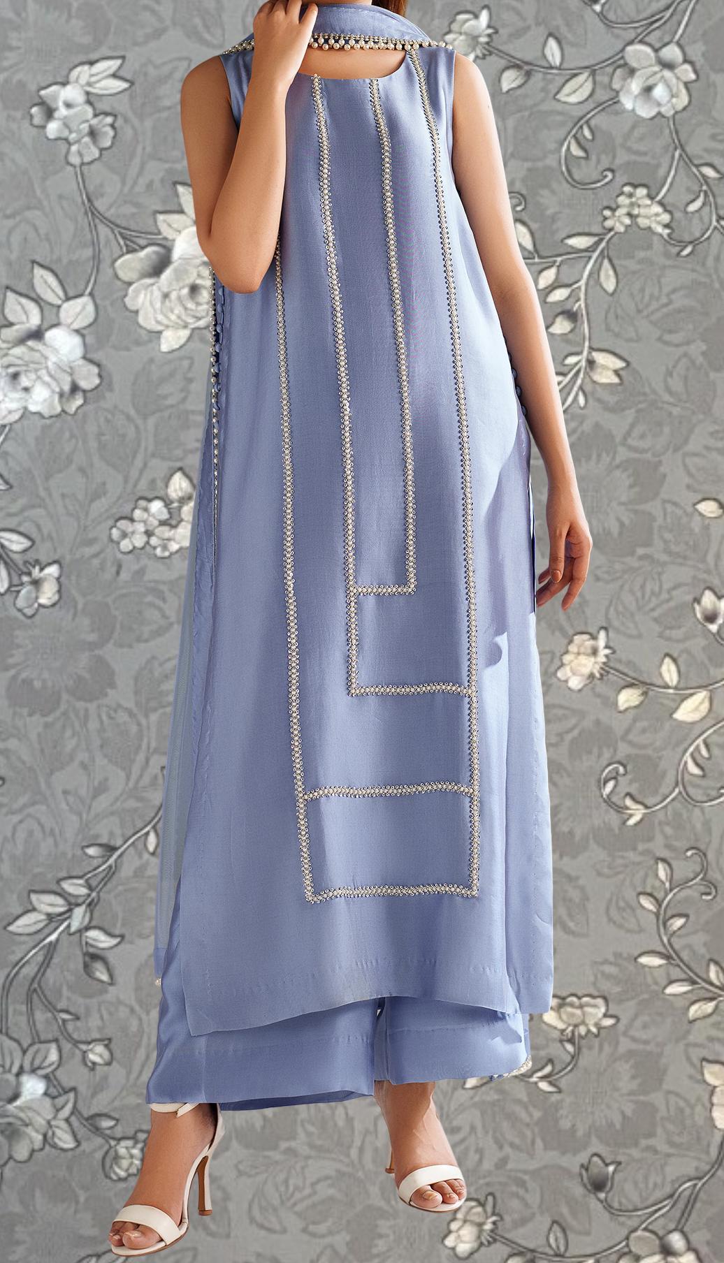 A woman wearing a long, sleeveless blue dress with a geometric pattern and white accents, standing against a gray floral background.