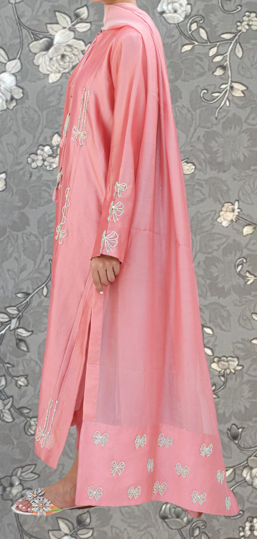 A person wearing a long pink dress with white floral embroidery stands against a gray floral background.