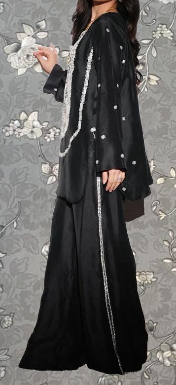 A woman wearing a long black dress with white accents, standing against a gray floral background.