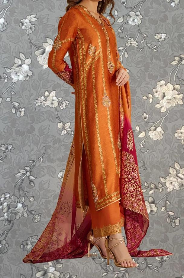A woman wearing an orange and red traditional Indian outfit with gold embroidery stands against a gray floral background.