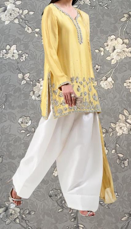 A woman wearing a yellow dress with gold embroidery and white pants, standing against a gray floral background.