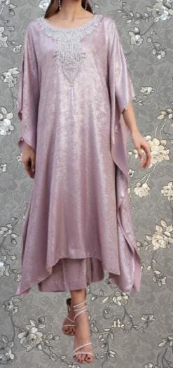 A woman wearing a long, flowing pink dress with a high neckline and intricate embroidery stands against a gray floral background.