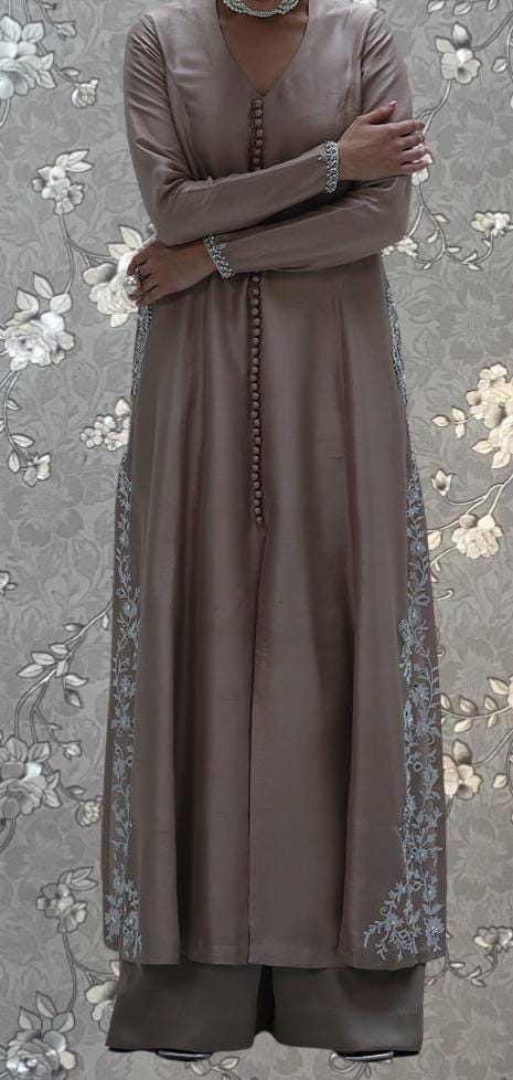 A woman wearing a long, gray dress with floral embroidery stands with her arms crossed against a floral background.