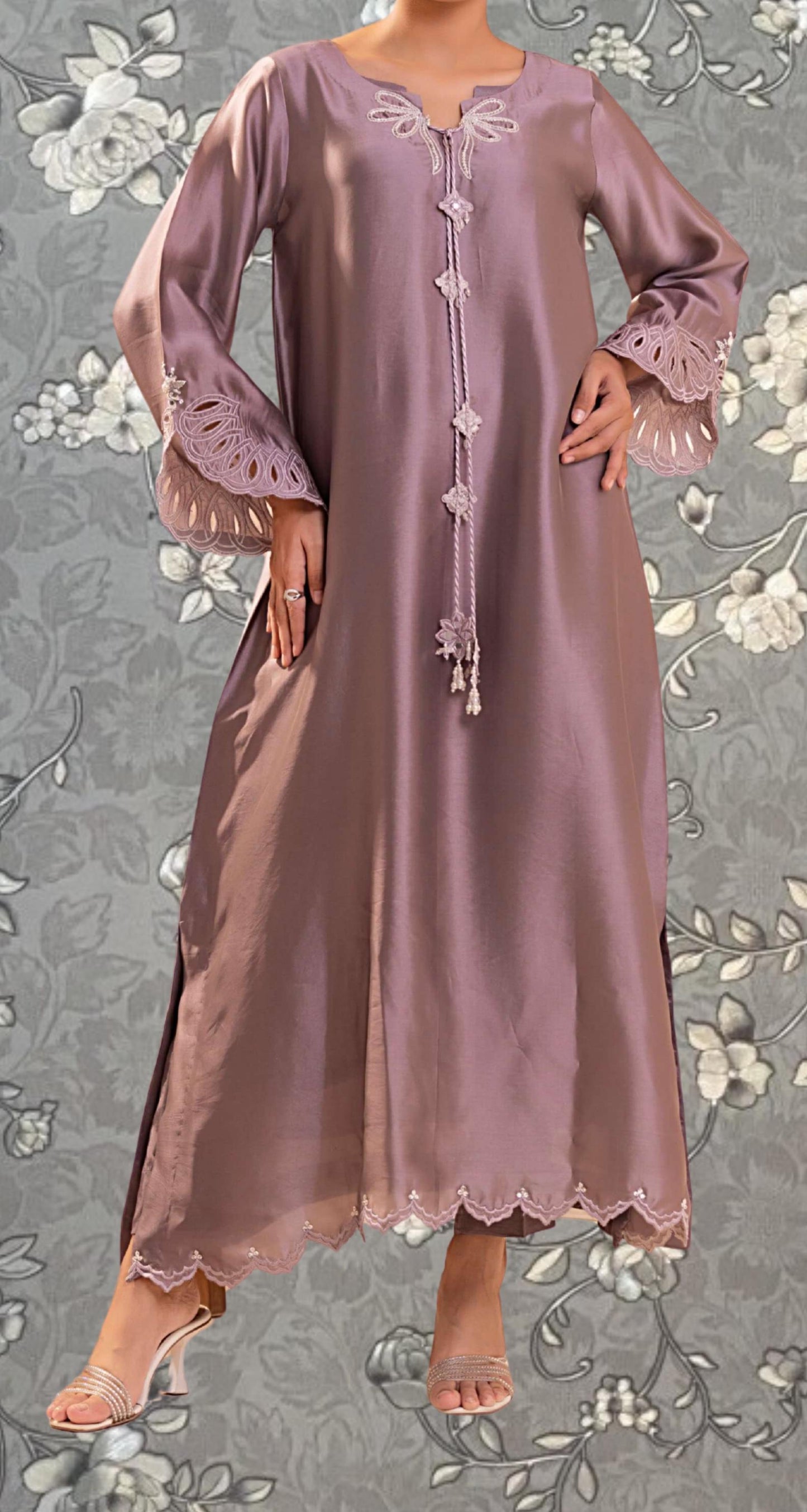 A woman wearing a long, purple dress with lace sleeves and a scalloped hem, standing against a gray floral background.