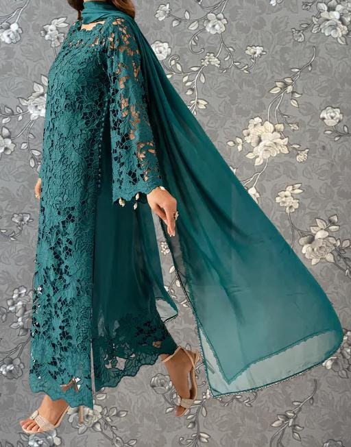 A woman wearing a long, green lace dress with a matching green shawl draped over her shoulders, standing against a gray floral background.