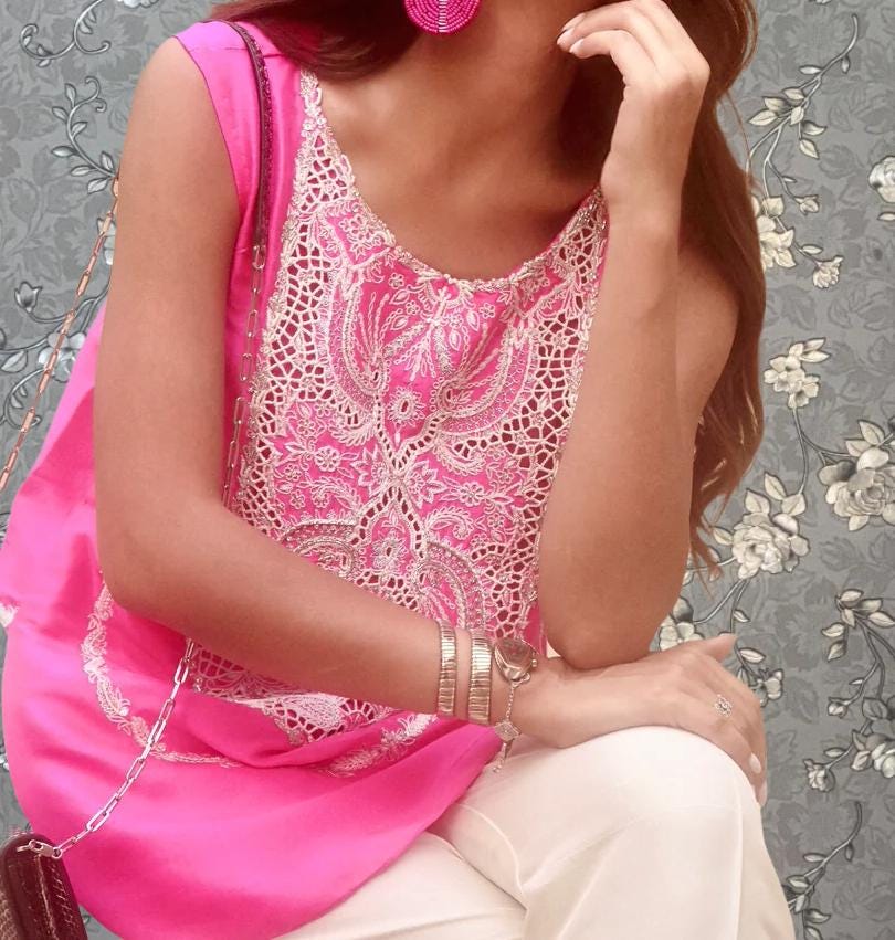 A woman wearing a pink and white patterned top and white pants is sitting with her hand on her chin, wearing a watch and bracelets.