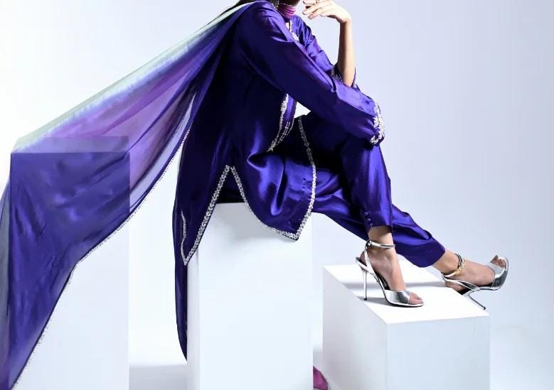 A person wearing a purple outfit with a flowing cape sits on a white pedestal against a plain white background.