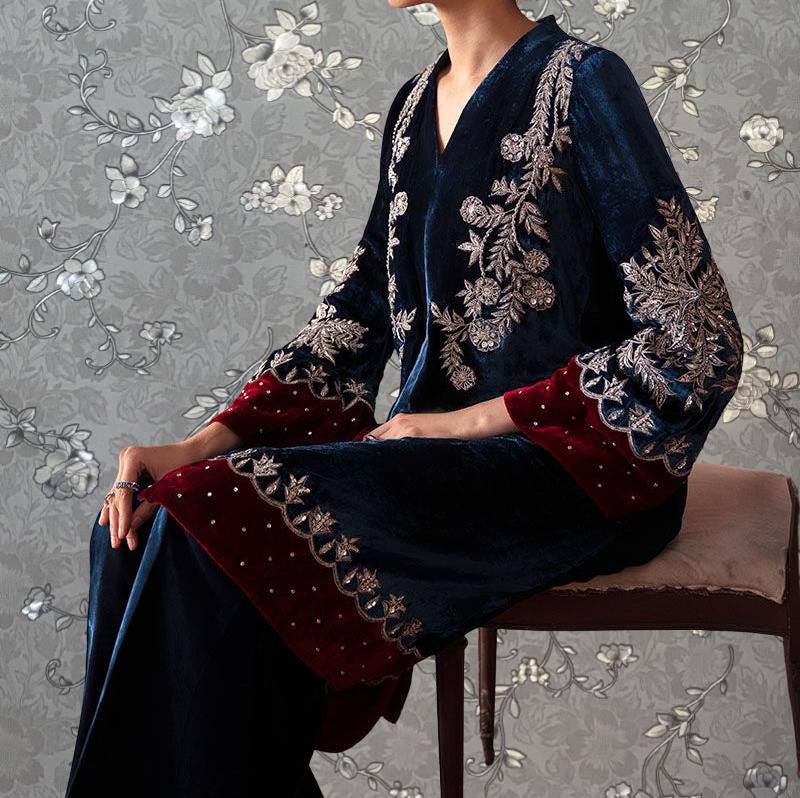A person wearing a blue velvet dress with red accents and intricate embroidery, sitting on a chair against a floral patterned background.