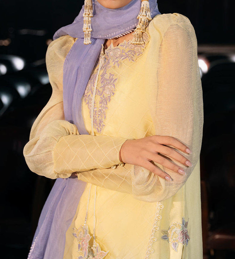 A woman wearing a yellow and purple dress with a purple scarf, posing with her arms crossed.