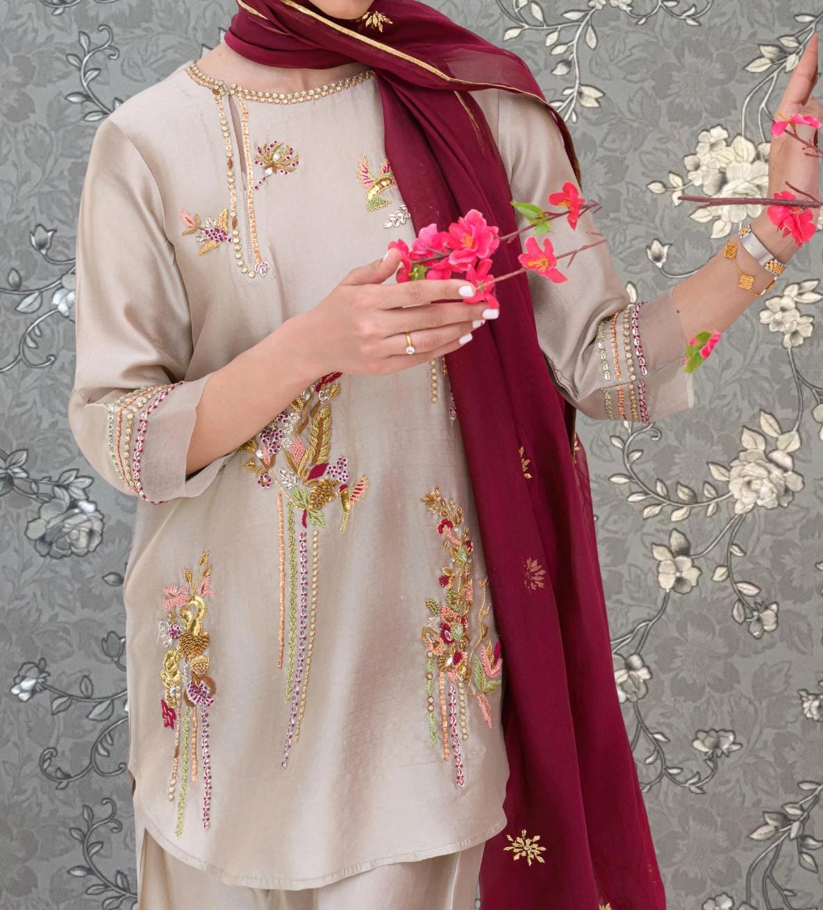 A person wearing a beige dress with intricate embroidery and holding a bouquet of pink flowers.