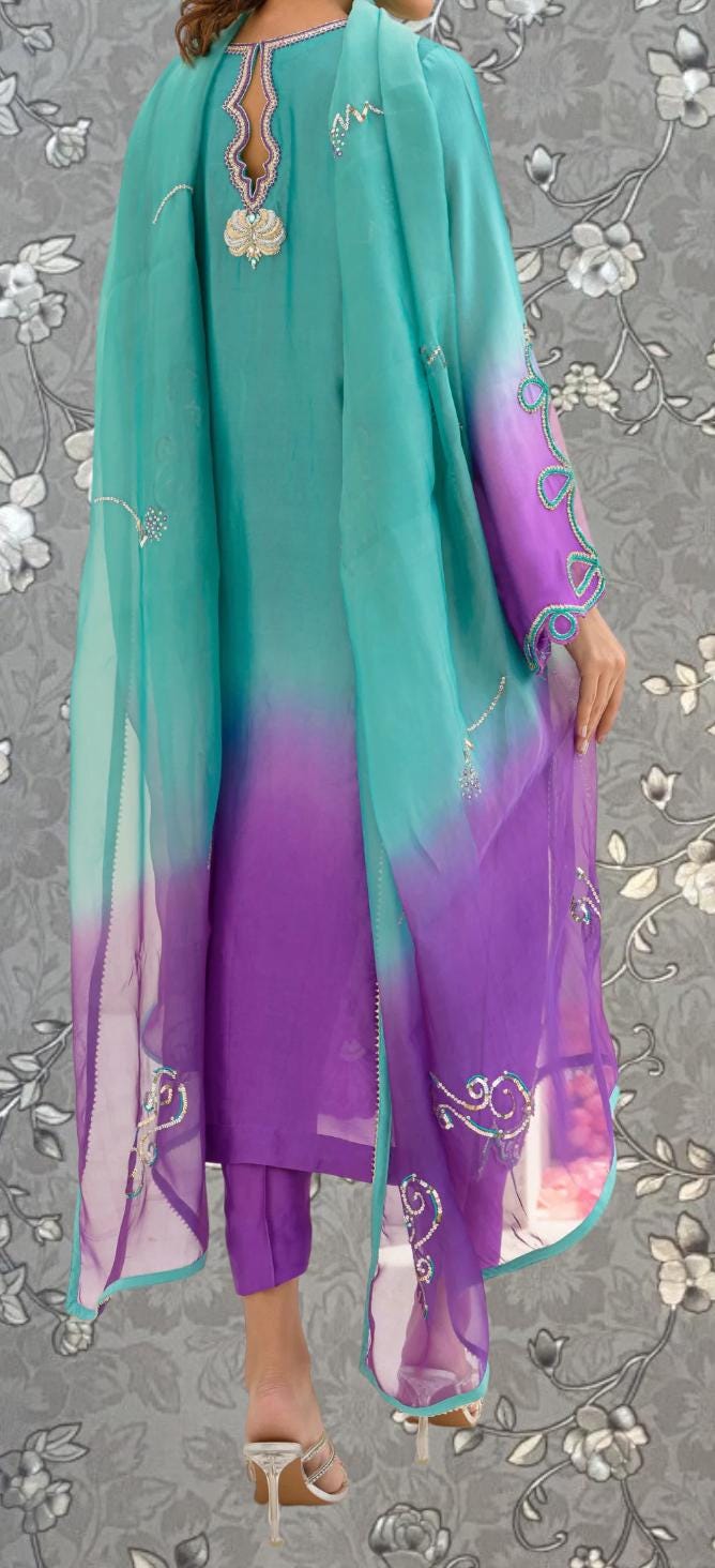 a person wearing a colorful, patterned dress with a turquoise and purple color scheme. The dress features a long, flowing cape and a floral pattern on the sleeves and hem. The person is standing against a gray floral background, which complements the vibrant colors of the dress.