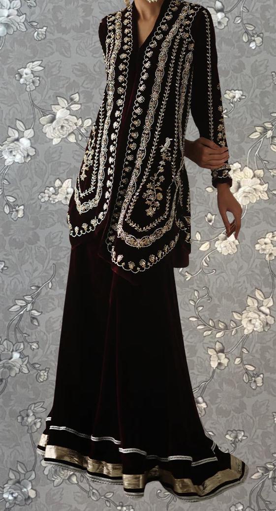 A woman wearing a long, dark dress with intricate embroidery and patterns, standing against a gray floral background.