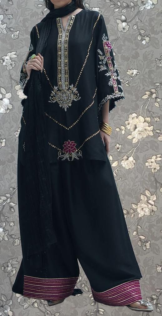 A person wearing a black dress with intricate embroidery and a matching black and pink skirt, standing against a floral background.