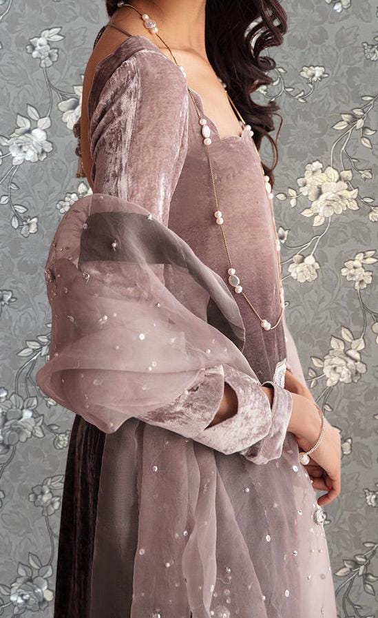 A woman wearing a long, pink velvet dress with a sheer, sheer-like fabric draped over her arm. She is also wearing a necklace with white pearls. The background features a floral pattern.