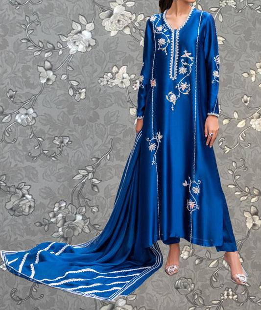 A woman wearing a long, blue dress with intricate embroidery stands against a gray floral background.