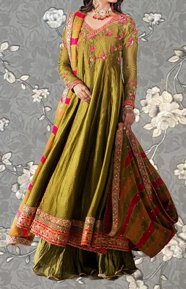 A woman wearing a long, green dress with intricate embroidery and a matching skirt, posing against a gray floral background.