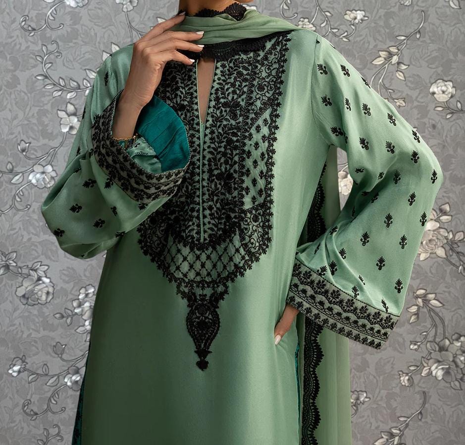 A person wearing a green dress with intricate black embroidery and patterns, posing against a gray floral background.
