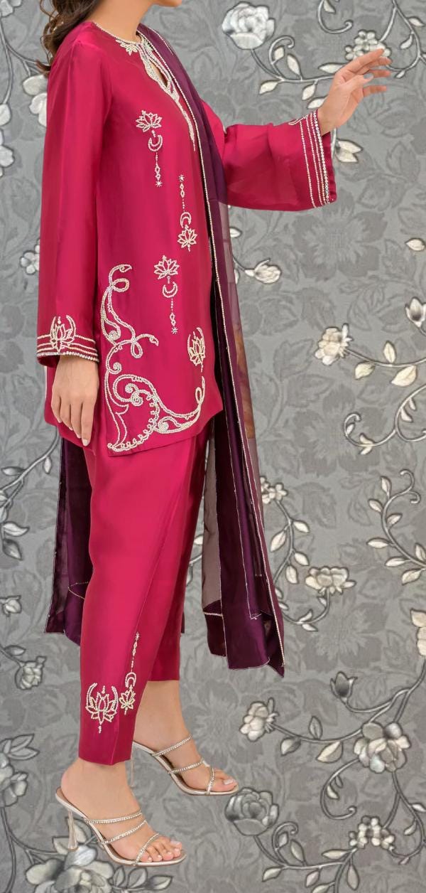 A woman wearing a vibrant red and purple outfit with intricate embroidery stands against a gray floral background.