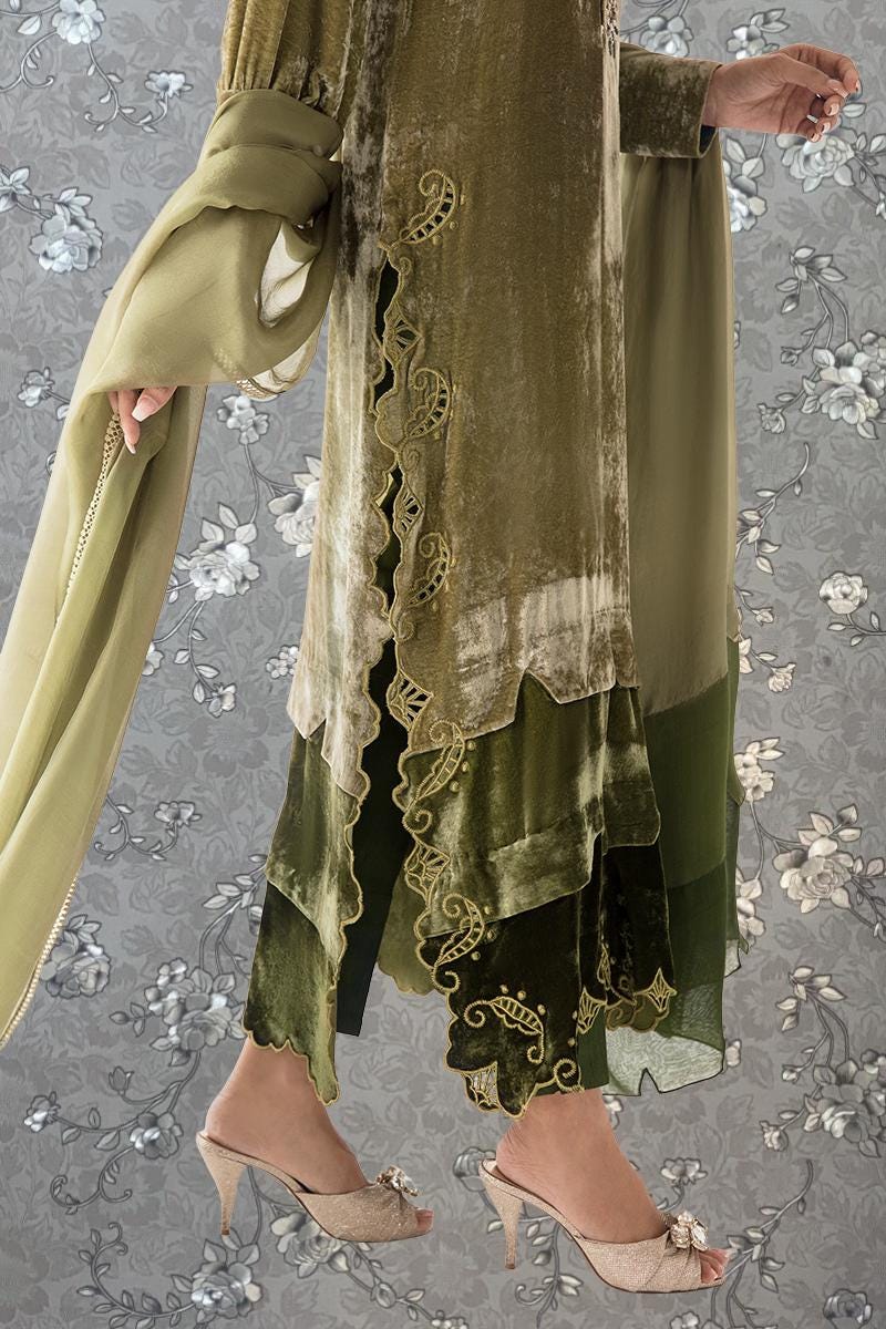 A person wearing a green velvet dress with intricate embroidery and a matching green shawl, standing against a gray floral background.