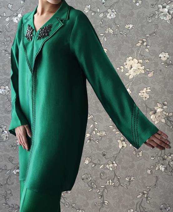 A person wearing a long green coat with floral embroidery on the collar and sleeves stands against a gray floral background.