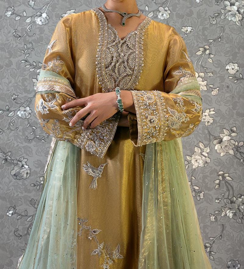A person wearing a gold and green embroidered dress with a matching gold and green shawl, standing against a gray floral background.