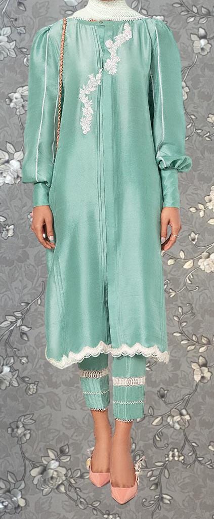 A person wearing a mint green dress with white lace trim and matching lace-trimmed pants, standing against a gray floral background.