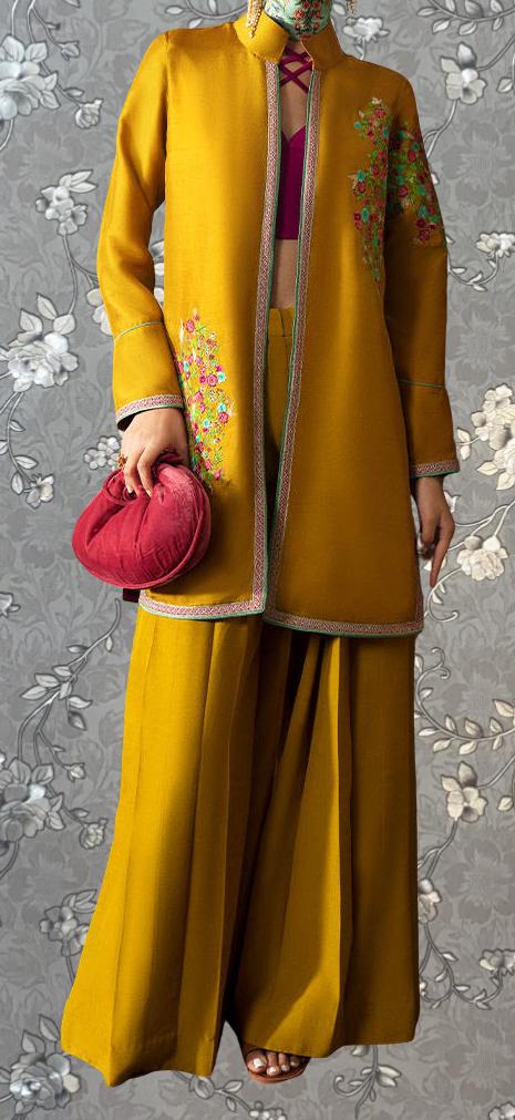 A person wearing a bright yellow jacket with floral embroidery and a matching skirt, holding a red purse, standing against a gray floral background.